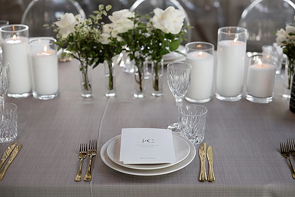 Reception tablescape with gray linen tablecloth, white rose centerpieces, pillar candles, gold flatware, and clear acrylic chairs in back