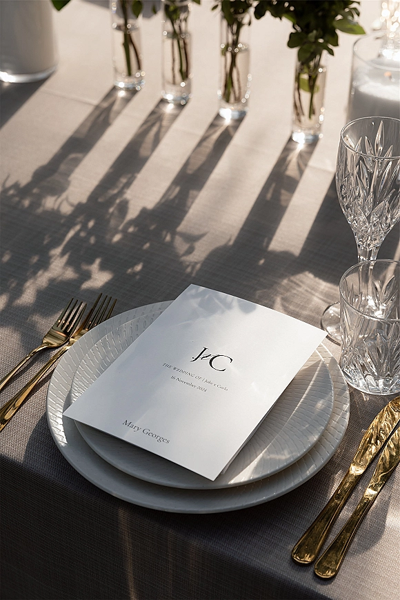 Wedding tablescape with wedding place setting of white plates, gold flatware, crystal goblets, and menu cards on gray linen in sunlight shadows