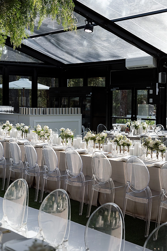 Reception tablescape with long banquet table wedding setup, white floral centerpieces and pillar candles in a glass conservatory overlooking trees