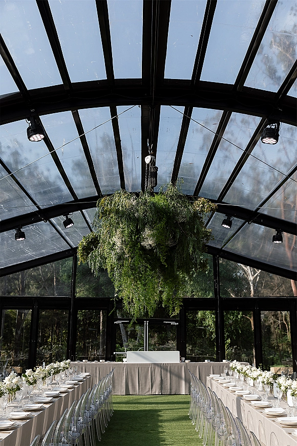 Reception tablescape with long banquet table setup, white linens and floral centerpieces under hanging greenery in a glass pavilion ceiling