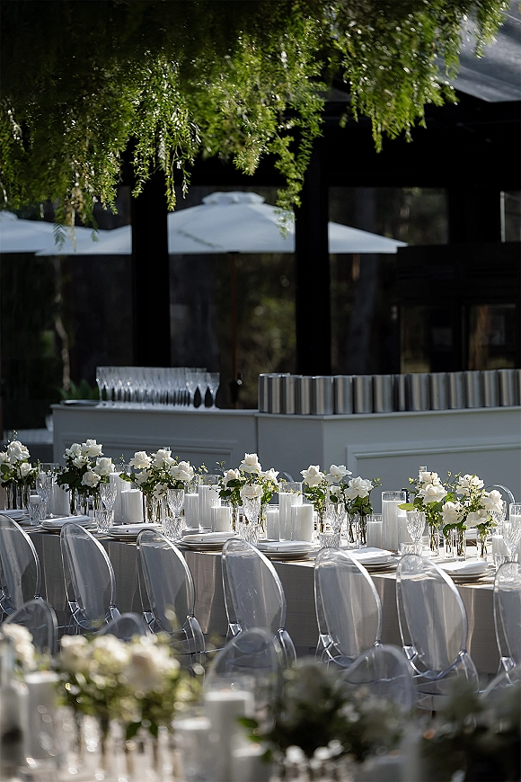 Reception tablescape with white rose centerpieces, bud vases and pillar candles on a long outdoor patio table under umbrellas and tree branches