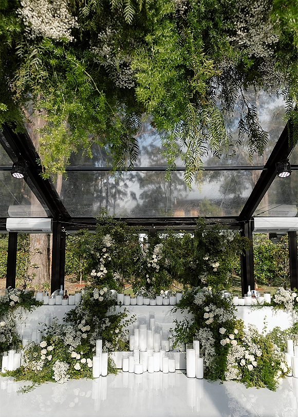 Wedding ceremony backdrop with greenery ceremony backdrop of white roses and baby's breath, pillar candles on white plinths in glass-ceiling venue