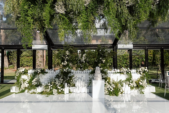 Wedding reception decor with a greenery wedding backdrop, white florals, pillar candles, cake display, and acrylic chairs in a glass pavilion on a lawn