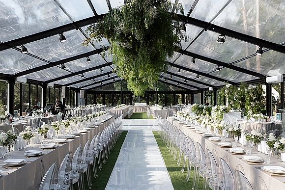 Reception tablescape with long banquet table setup, beige linens, clear acrylic chairs, white florals, and hanging greenery in a glass tent by forest trees