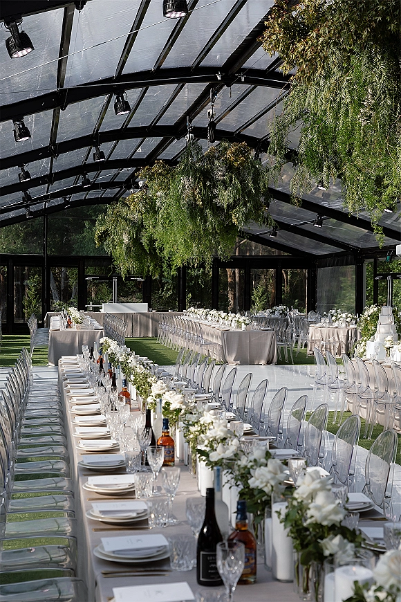 Reception tablescape with long banquet table setup, white florals and greenery garlands, ghost chairs, candles, and tiered cake under glass roof