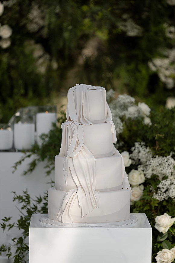 Wedding cake with modern wedding cake styling, tiered white fondant draping on a pedestal table with white roses, candles, and greenery backdrop