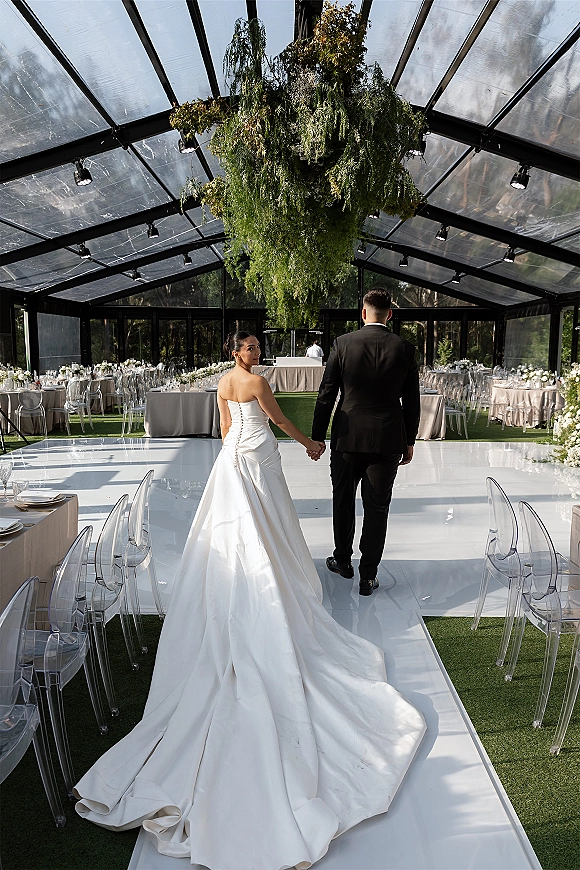 Couple portrait of bride and groom holding hands on a white aisle runner, bride in a strapless gown with long train in a glass greenhouse tent