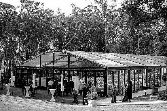 Wedding reception venue with a clear marquee tent, cocktail tables and patio umbrellas, set by a garden lawn walkway and trees at night