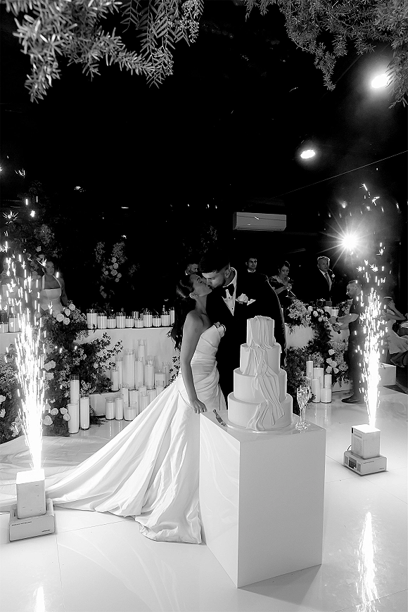 Wedding cake cutting as bride in a satin gown and groom in a black tux slice a tiered cake with sparkler fountain accents on a stage