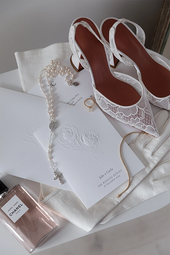 Wedding flat lay featuring a wedding invitation suite with lace slingback heels, pearl rosary, rings, gloves, and perfume on a white tabletop in window light