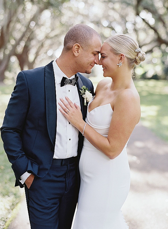 Couple portrait of bride and groom close up, touching foreheads as her hand rests on his chest, tree-lined path behind them