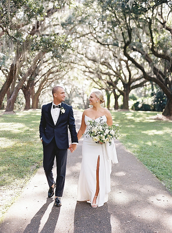 Couple portrait of bride and groom walking hand in hand, bride holding greenery bouquet, along a sunlit oak-lined path with moss