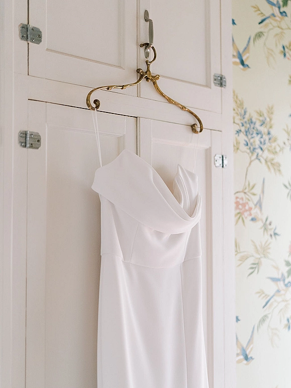 Wedding dress hanging on a dress hanger against white wardrobe doors and floral wallpaper, showcasing minimalist wedding dress clean lines