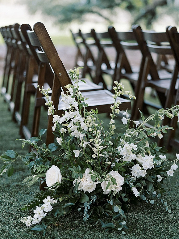 Ceremony aisle decor with outdoor ceremony chairs, wooden folding chairs lined by white roses and greenery garland on a grass lawn