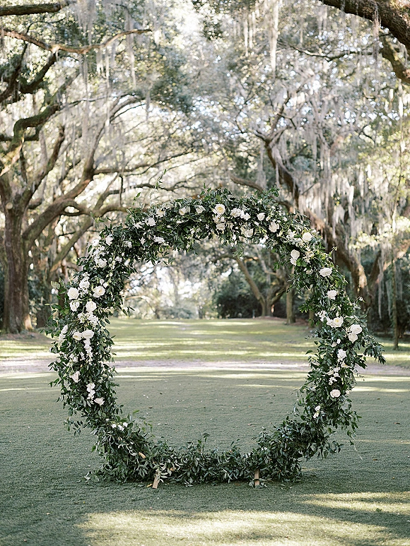 Wedding ceremony arch with a circular wedding arch design, greenery garland and white flowers, set on a lawn beneath oak trees and moss