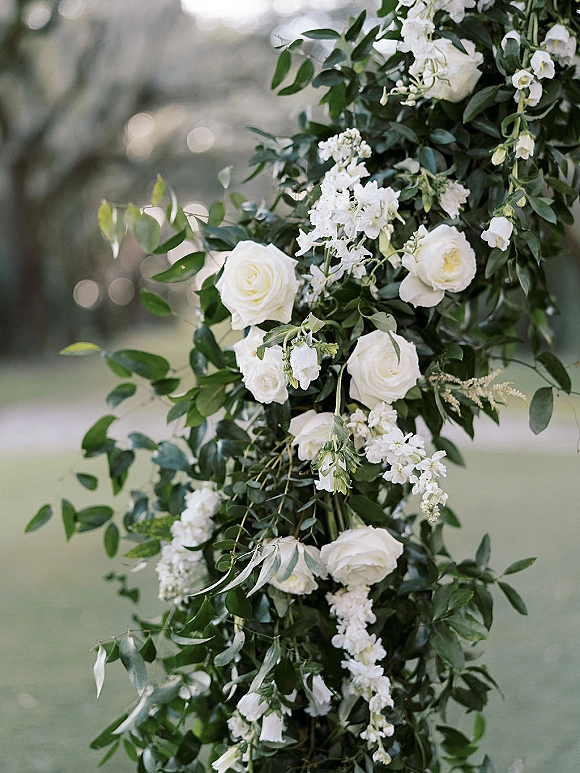 Wedding floral arrangement of white rose wedding flowers with cascading greenery on a ceremony pillar in a garden lawn setting