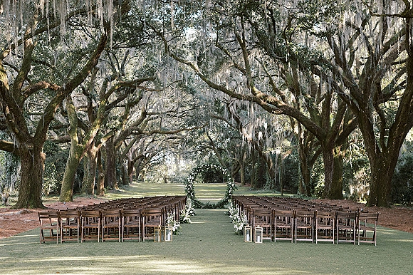 Ceremony setup for an outdoor wedding ceremony with wood chairs flanking a lantern-lined aisle under oak trees with hanging moss