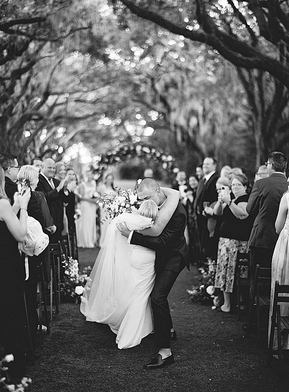Wedding kiss as newlyweds kissing aisle, bride in strapless gown with veil and bouquet, guests seated under string lights and trees