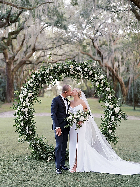 Wedding kiss as bride and groom embrace under a greenery floral arch with white flowers, cathedral veil flowing beneath oak trees and moss