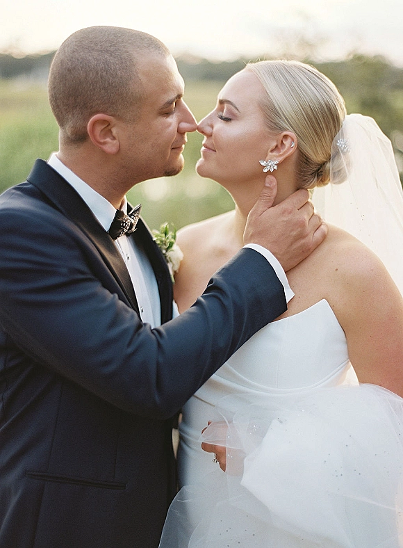Wedding kiss portrait of bride and groom kiss as he holds her face, veil flowing in golden sunset light over an open field