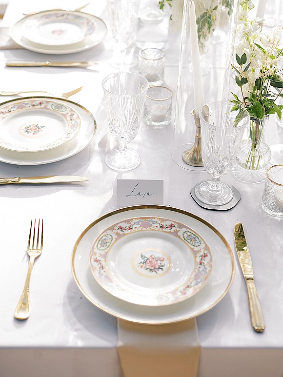 Reception tablescape with wedding place setting on white tablecloth, floral china, gold flatware, crystal goblets, candles, and white flowers in bud vases.