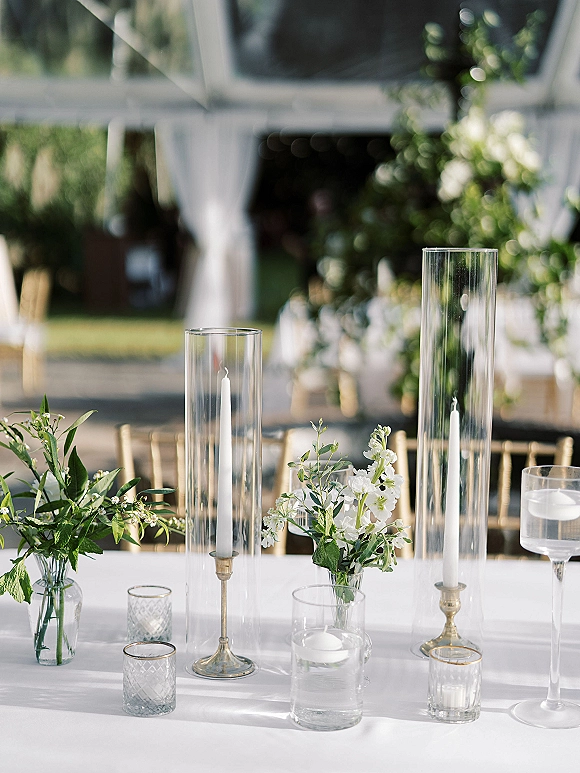 Reception tablescape with wedding table centerpiece featuring tall hurricane vases, taper candles, and white flowers under a draped outdoor tent