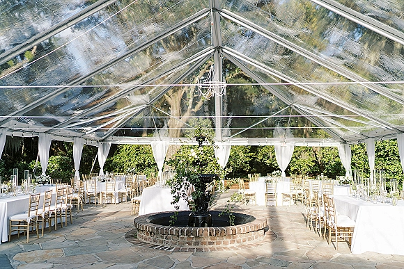 Tent reception decor with a clear top tent, white draping, chandeliers and string lights over white-and-gold tables on a stone patio