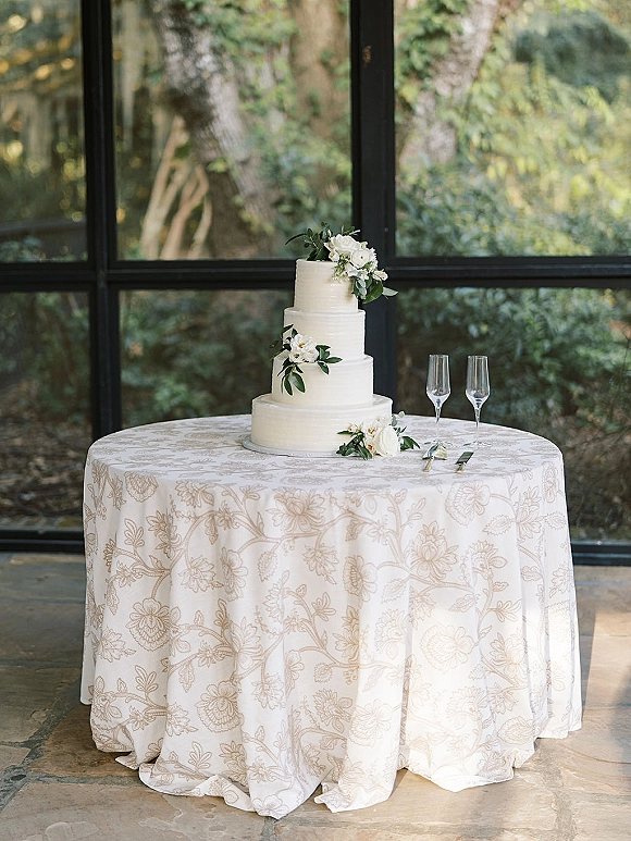 Wedding cake with a three tier wedding cake design, white flowers and greenery on a stand beside champagne flutes by garden-lit windows