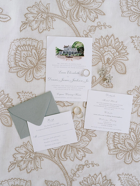 Invitation suite with watercolor venue invitation, sage green envelopes, RSVP and details cards, rings and earrings on neutral linen backdrop