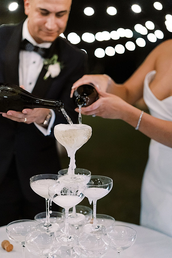 Champagne tower pour as bride and groom tip bottles over coupe glasses, foam cascading under string lights in the night sky