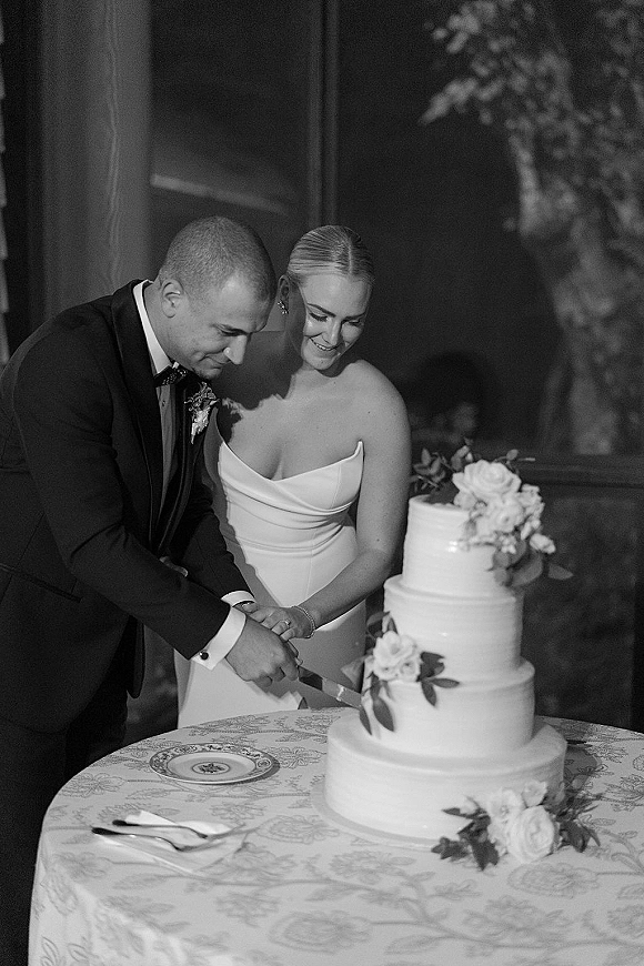 Wedding cake cutting as bride in a strapless gown and groom in tux hold a knife by a floral-topped cake on a lace tablecloth