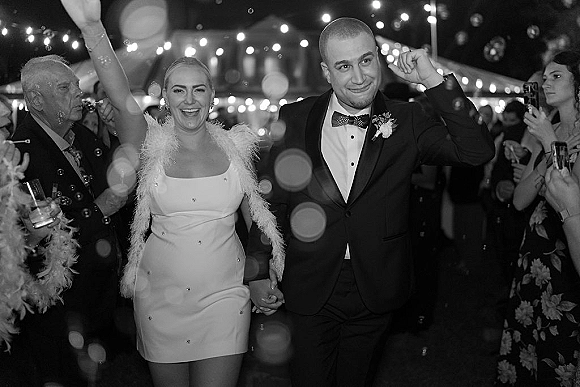 Wedding reception entrance as newlyweds make a grand entrance under bistro lights, bubbles swirling while guests cheer outdoors at night