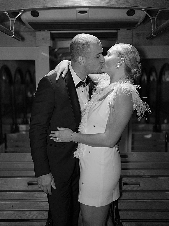 Wedding kiss as bride in sleeveless short dress and feather wrap embraces groom in bow tie tuxedo at a bar with glass racks