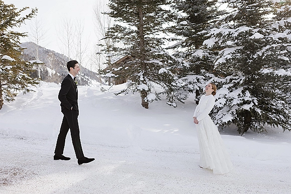 First look moment as bride in a wedding dress and bridal coat faces groom in a tuxedo on a snowy mountain path with evergreens