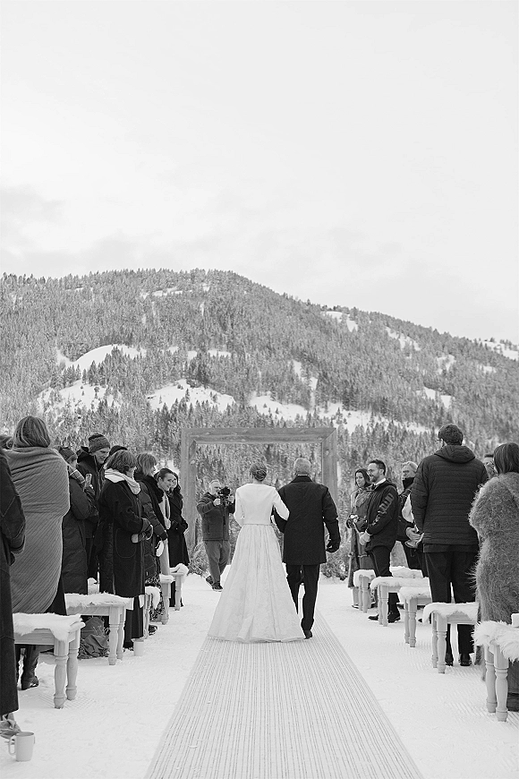 Wedding processional with bride walking down aisle in a long sleeve jacket and gown toward a wooden arch, snowy pines and mountains behind