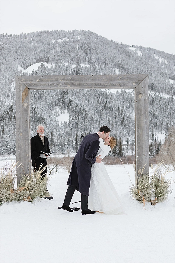 Ceremony kiss at an outdoor winter ceremony under a rustic wooden arch with greenery, pine trees and snowy mountains behind