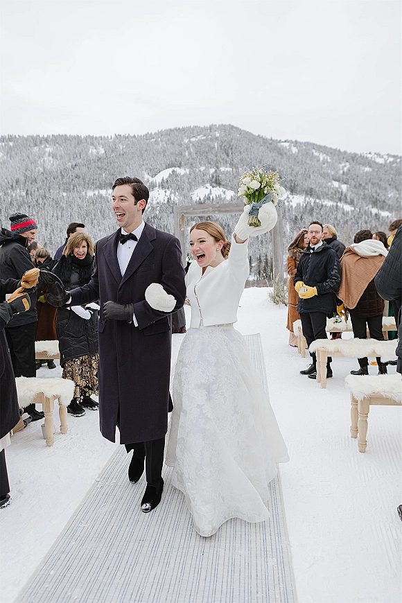 Wedding recessional as bride and groom walk the aisle in winter coats, bouquet raised, with snowy mountains and evergreen trees behind them