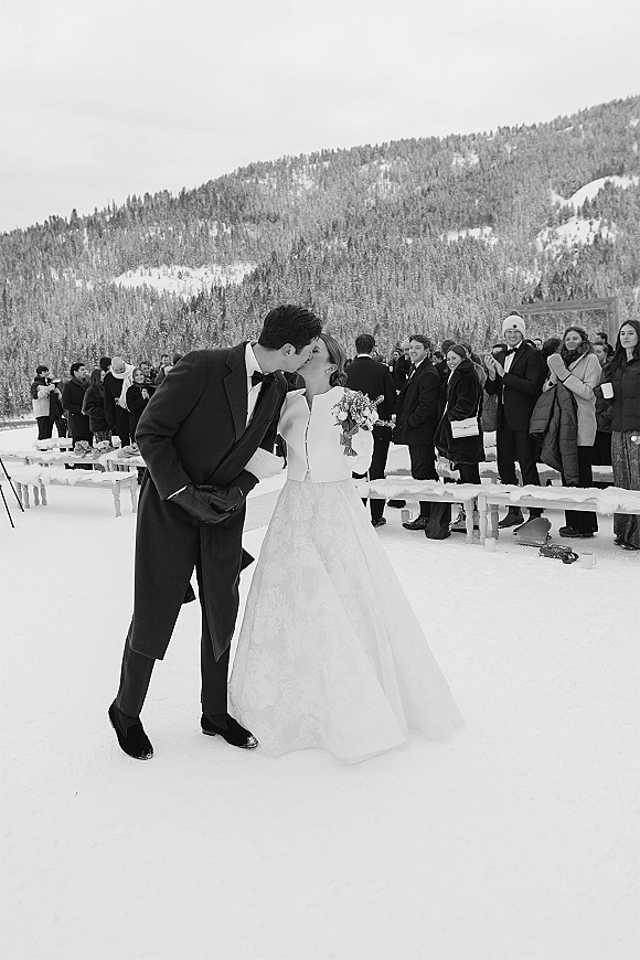 Wedding kiss at an outdoor winter wedding, bride in long sleeve dress holding a bouquet as guests watch amid snow and pine mountains