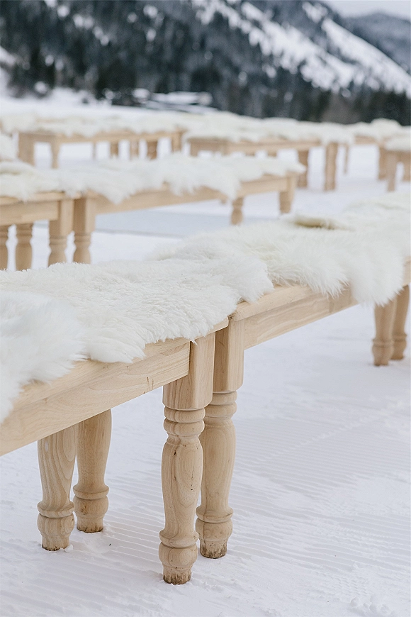 Ceremony seating with outdoor wedding benches in snow, natural wood benches topped with white fur throws amid evergreens and mountains