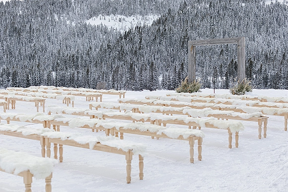 Ceremony setup for an outdoor winter ceremony with wooden arch and greenery, fur-covered benches, and a snowy pine forest mountainside backdrop