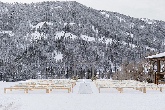 Outdoor ceremony setup with a white aisle runner leading to a square arch with dried grasses, wood benches on snow by pines and cabin
