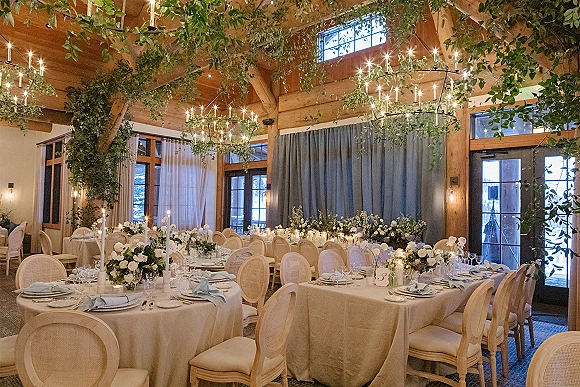 Reception tablescape with wedding reception table decor featuring white and greenery centerpieces, taper candles, and blue napkins under chandeliers and string lights