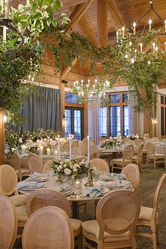 Reception tablescape with round wedding tables set with taper candles and greenery garlands beneath chandeliers in a wood-beam room with windows