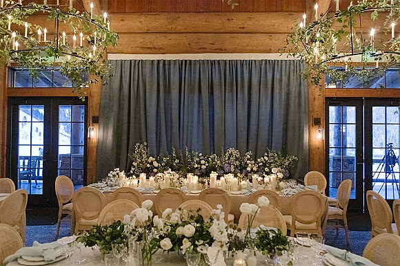 Reception sweetheart table with candlelit clusters and lush greenery garland beneath wooden beams, draped backdrop, and French doors with snow view