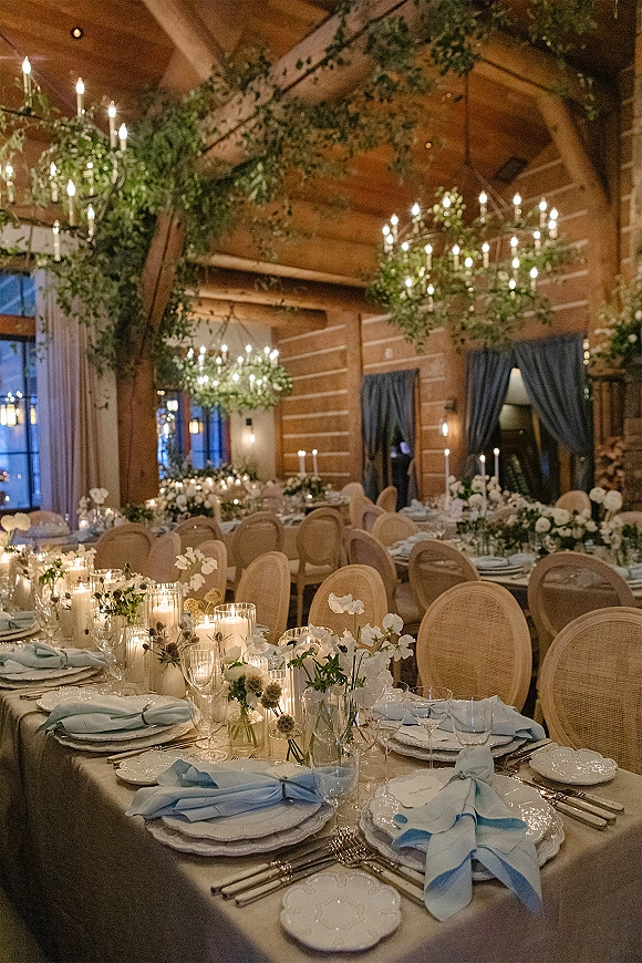 Reception tablescape with a candlelit reception table, taper candles and white floral centerpieces beneath chandeliers in a wood-beam hall