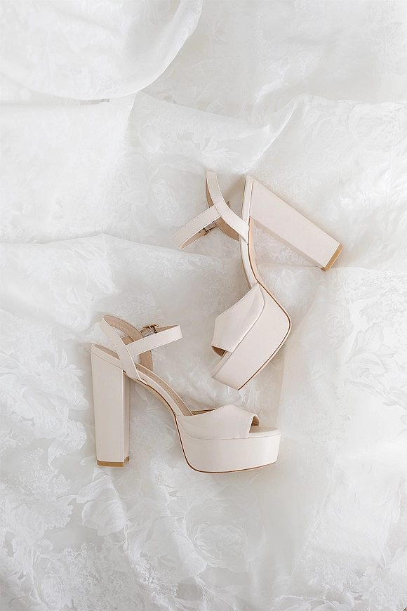Bridal shoes ivory bridal heels with ankle strap and block heel, arranged on white floral fabric for a clean wedding detail flat lay
