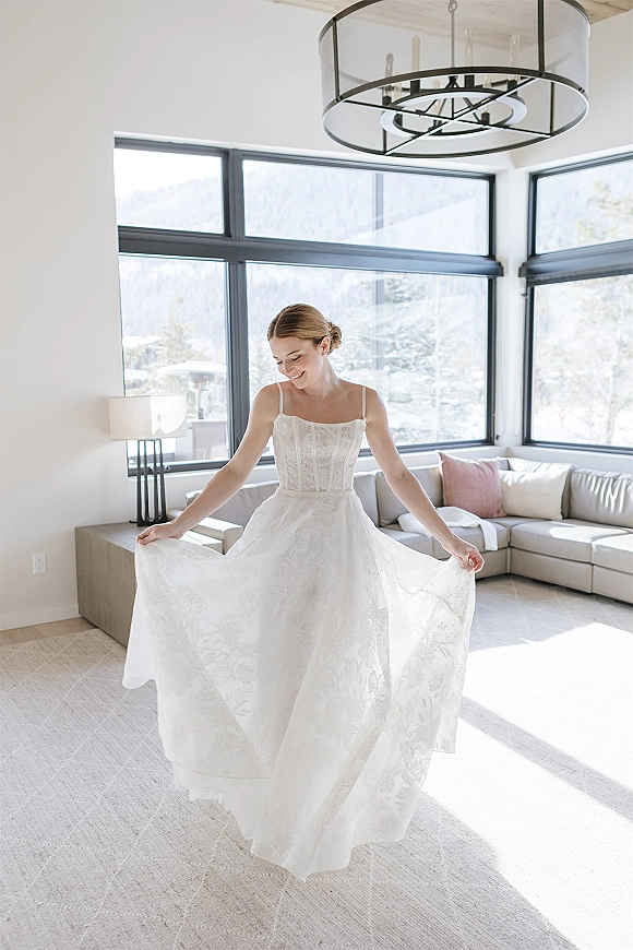 Bridal portrait of a bride twirling her full skirt in a lace bodice gown by large windows, with snowy mountains beyond the suite