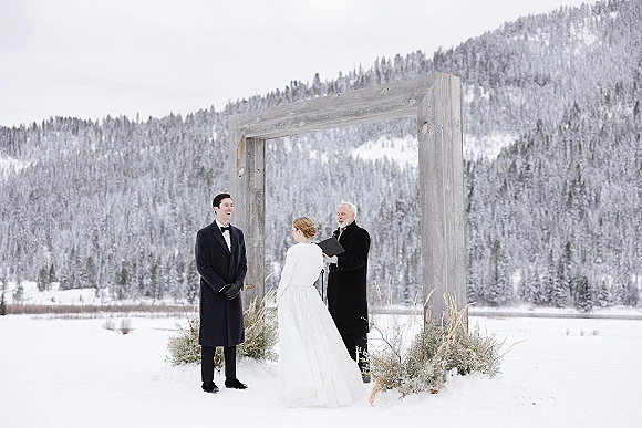 Wedding ceremony with bride and groom at a greenery arch as the officiant reads, set against snowy mountains and pine trees