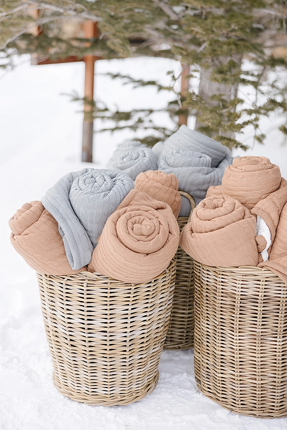 Wedding guest blankets in a wedding blanket basket, with knit throws and rolled blankets beside snow, evergreens, and a wooden structure