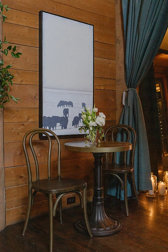 Cocktail table decor with a bud vase centerpiece of white flowers and greenery, set by a wood plank wall with candles and bistro chairs